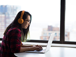 A woman wearing headphones using a laptop in front of a window.
