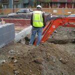 A man in a safety vest standing in a pile of excavated dirt on a construction site watching the excavator.